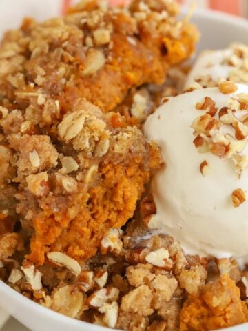 Closeup of a pumpkin crisp in a white bowl topped with vanilla ice cream and chopped pecans.