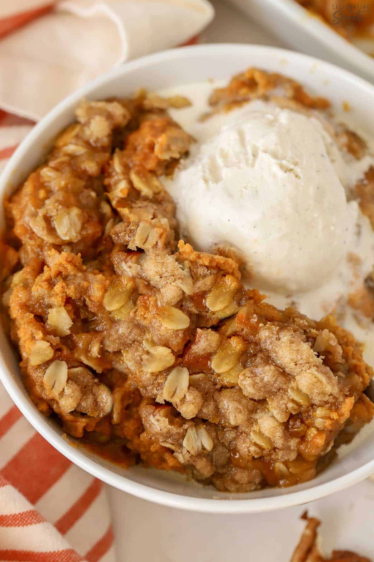 Closeup of a pumpkin crisp in a white bowl topped with ice cream. Next to the bowl is an orange and white striped towel.