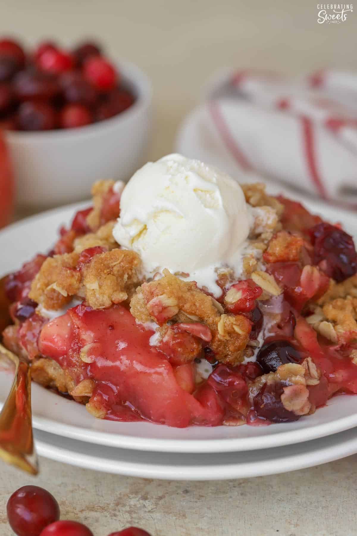 Apple cranberry crisp topped with vanilla ice cream on a white plate next to a gold spoon.