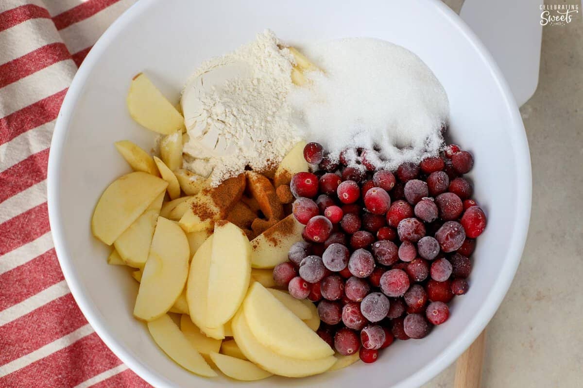 Apple slices, cranberries, sugar, flour, and cinnamon in a large white bowl next to a red striped napkin.