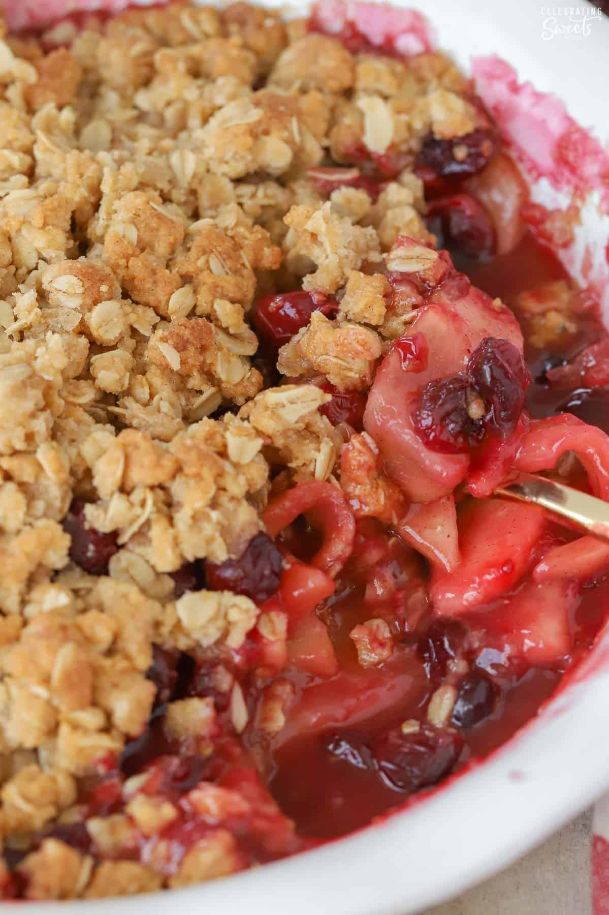 Apple cranberry crisp in a white baking dish with a gold spoon.