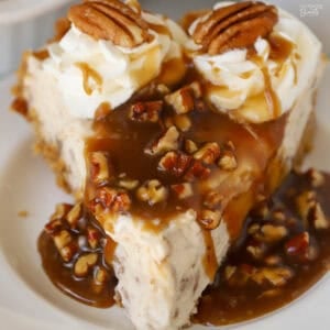 A slice of pecan pie cheesecake covered in pecan sauce and topped with whipped cream.
