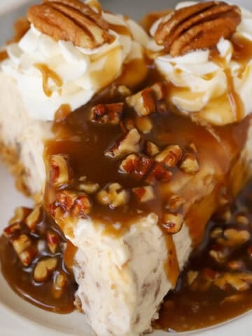 A slice of pecan pie cheesecake covered in pecan sauce and topped with whipped cream.