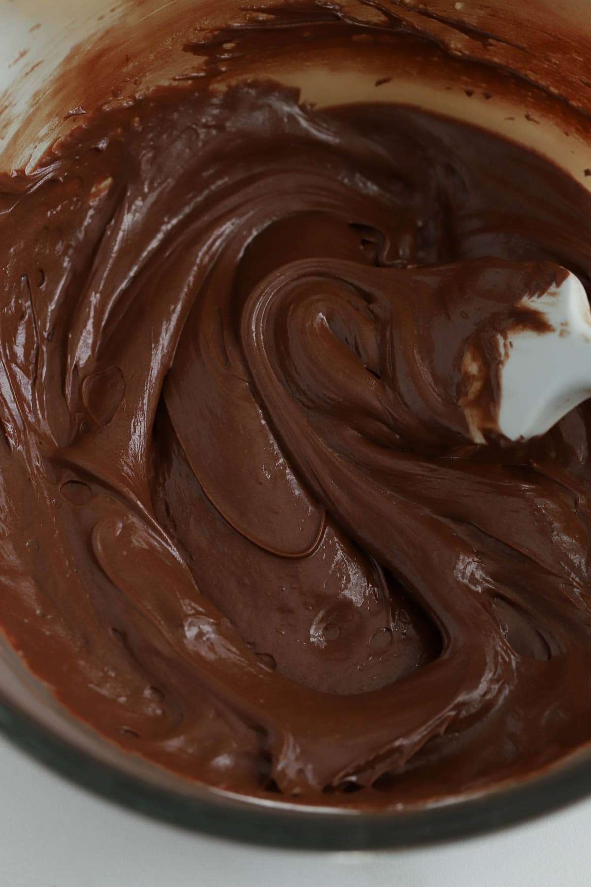 Creamy chocolate frosting in a glass bowl with a rubber spatula.