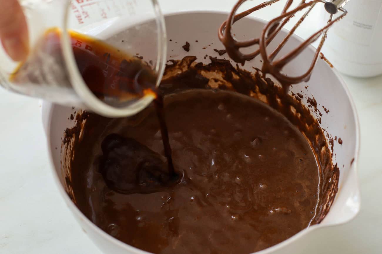 Coffee being poured into chocolate cake batter in a white bowl.