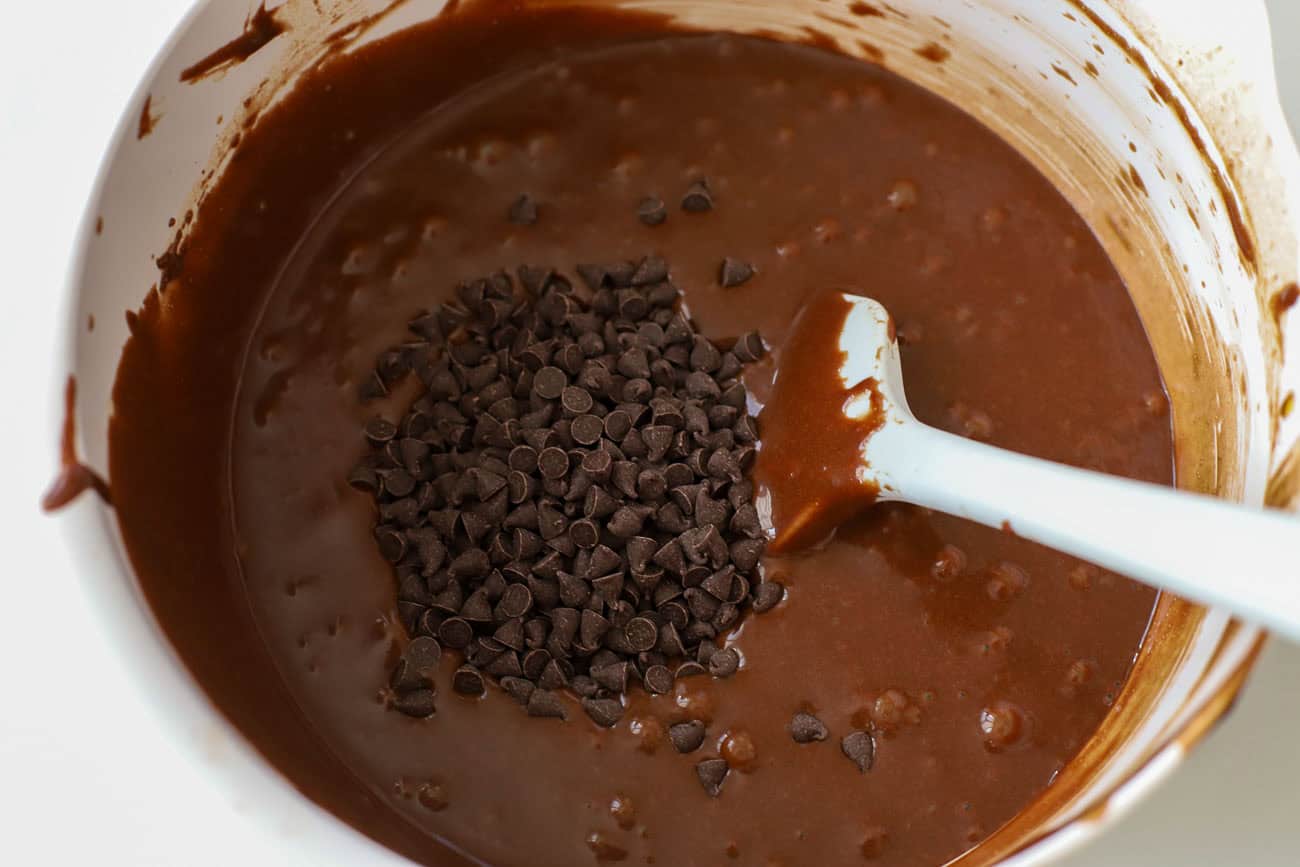 Chocolate cake batter in a white bowl topped with mini chocolate chips and a rubber spatula in the bowl.