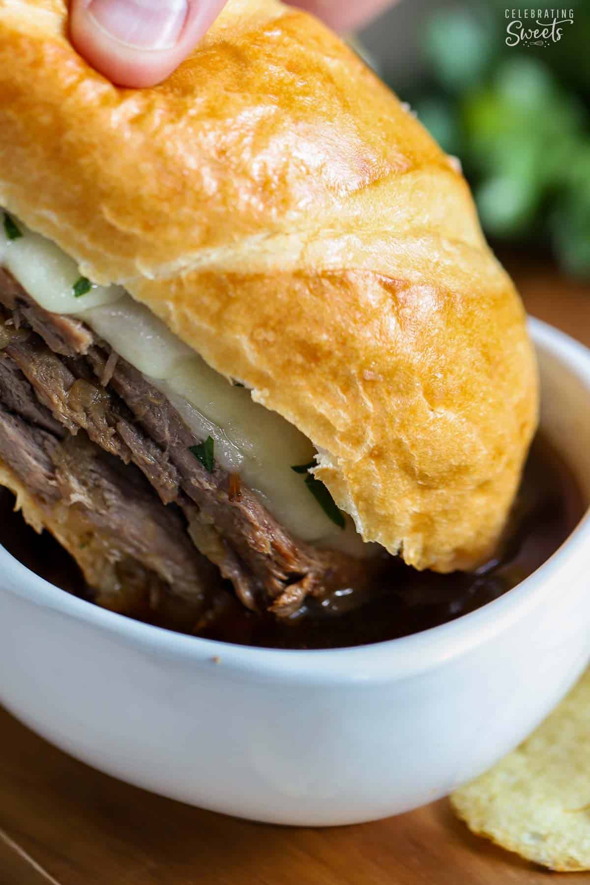 French dip sandwich being dipped in au jus in a white bowl on a wooden cutting board.