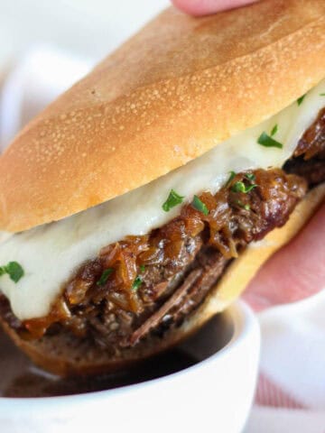 French dip sandwich being dipped in au jus in a white bowl.