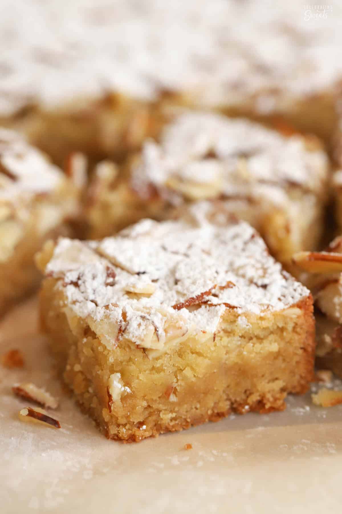 Closeup of an almond blondie topped with sliced almonds and powdered sugar on a piece of parchment paper.