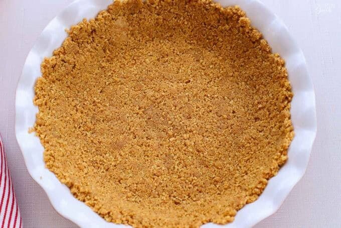 Graham cracker crust in a white pie plate next to a red and white striped napkin.