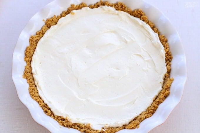 Cream cheese filling in a graham cracker crust in a white and red pie plate.