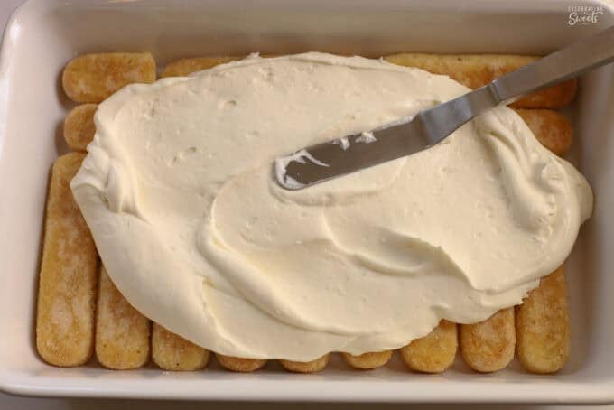 Whipped cream being spread onto a layer of ladyfingers in a baking dish.