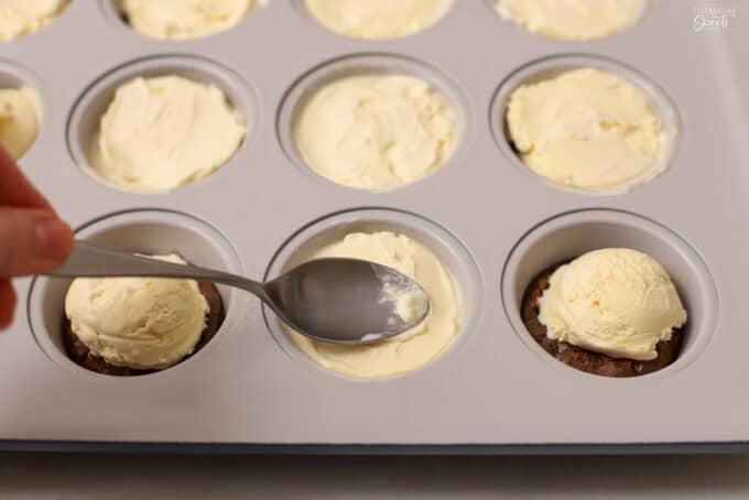 Ice cream being spooned on top of brownies in a muffin pan.