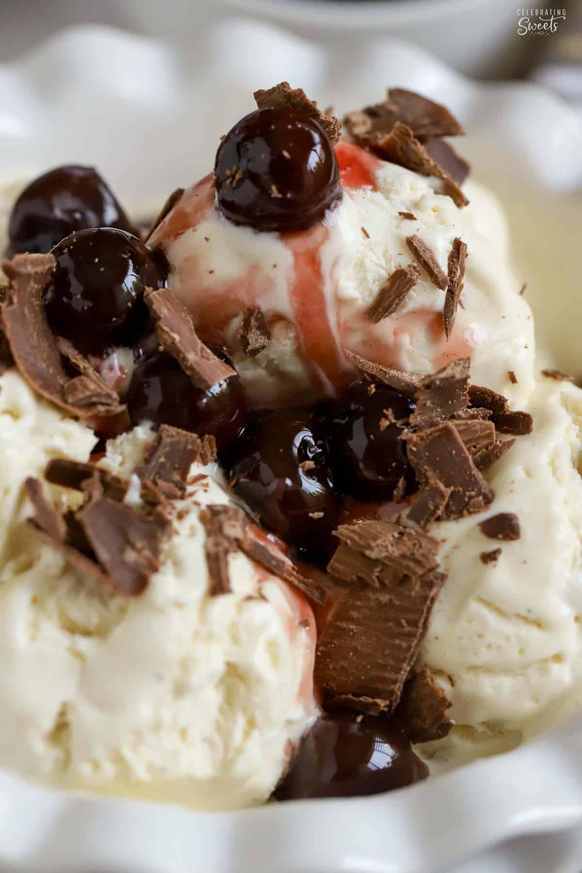 Scoops of vanilla ice cream in a white bowl topped with chocolate shavings and cherries.