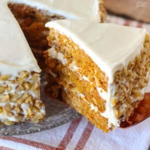 Slice of three layer pumpkin carrot cake topped with white frosting and walnuts on a glass plate.