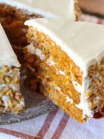 Slice of three layer pumpkin carrot cake topped with white frosting and walnuts on a glass plate.