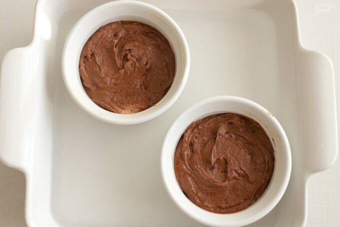 Brownie batter in two round ramekins sitting in a white square baking dish.