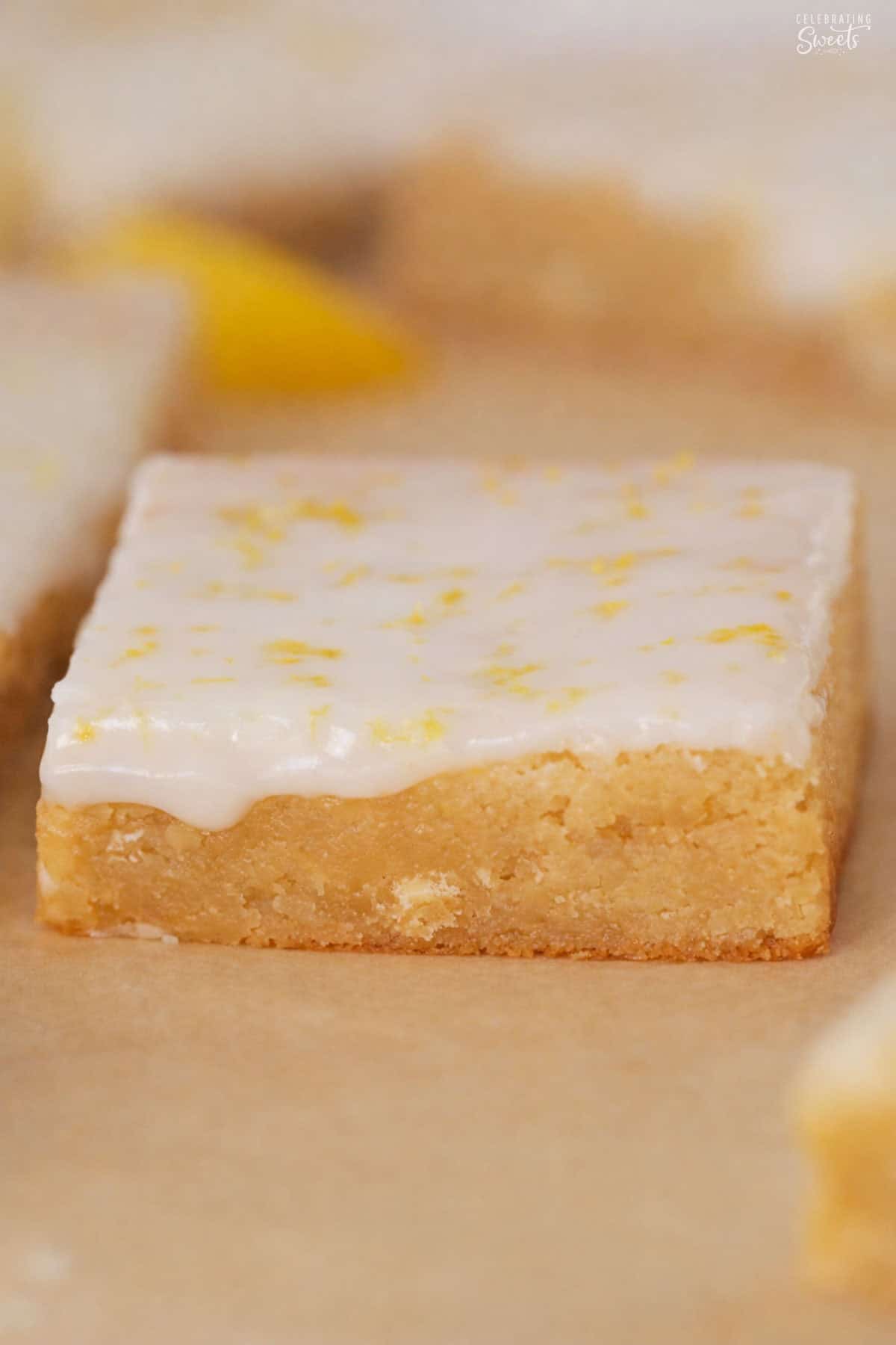 A slice of lemon brownie topped with white icing on parchment paper.