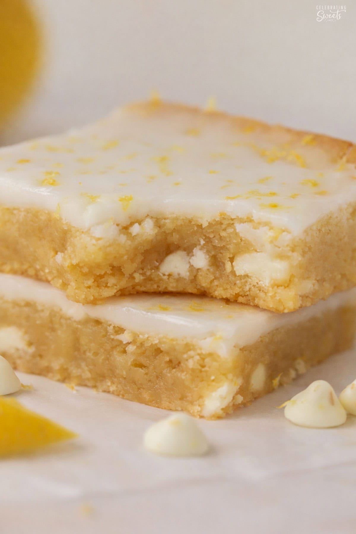 Stack of two lemon brownies on a piece of parchment paper next to a lemon slice and white chocolate chips.