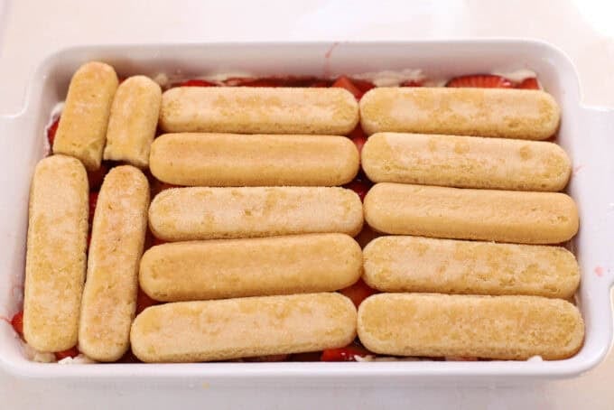 Ladyfingers on top of strawberries in a white rectangular baking dish.