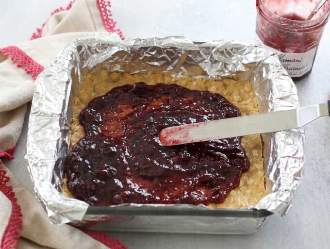 Jam being spread on oatmeal cookie bars in a foil-lined baking dish.
