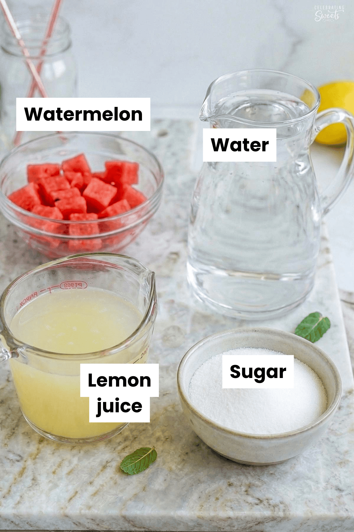 Lemon juice, sugar, a pitcher of water, and a bowl of cubed watermelon on a marble board.