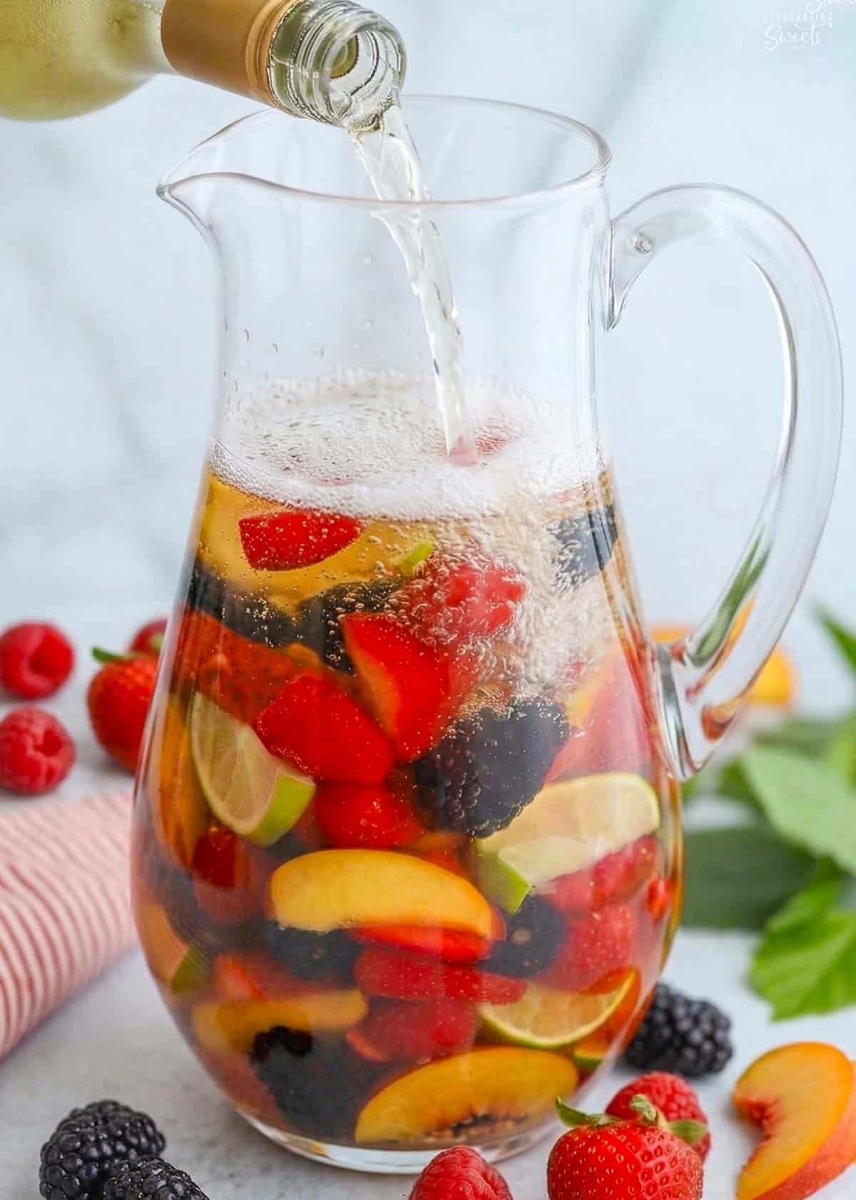 White wine being poured into a glass pitcher filled with fresh fruit.