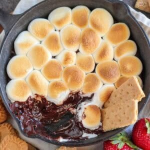 S'mores dip in a cast iron skillet next to graham crackers and strawberries.