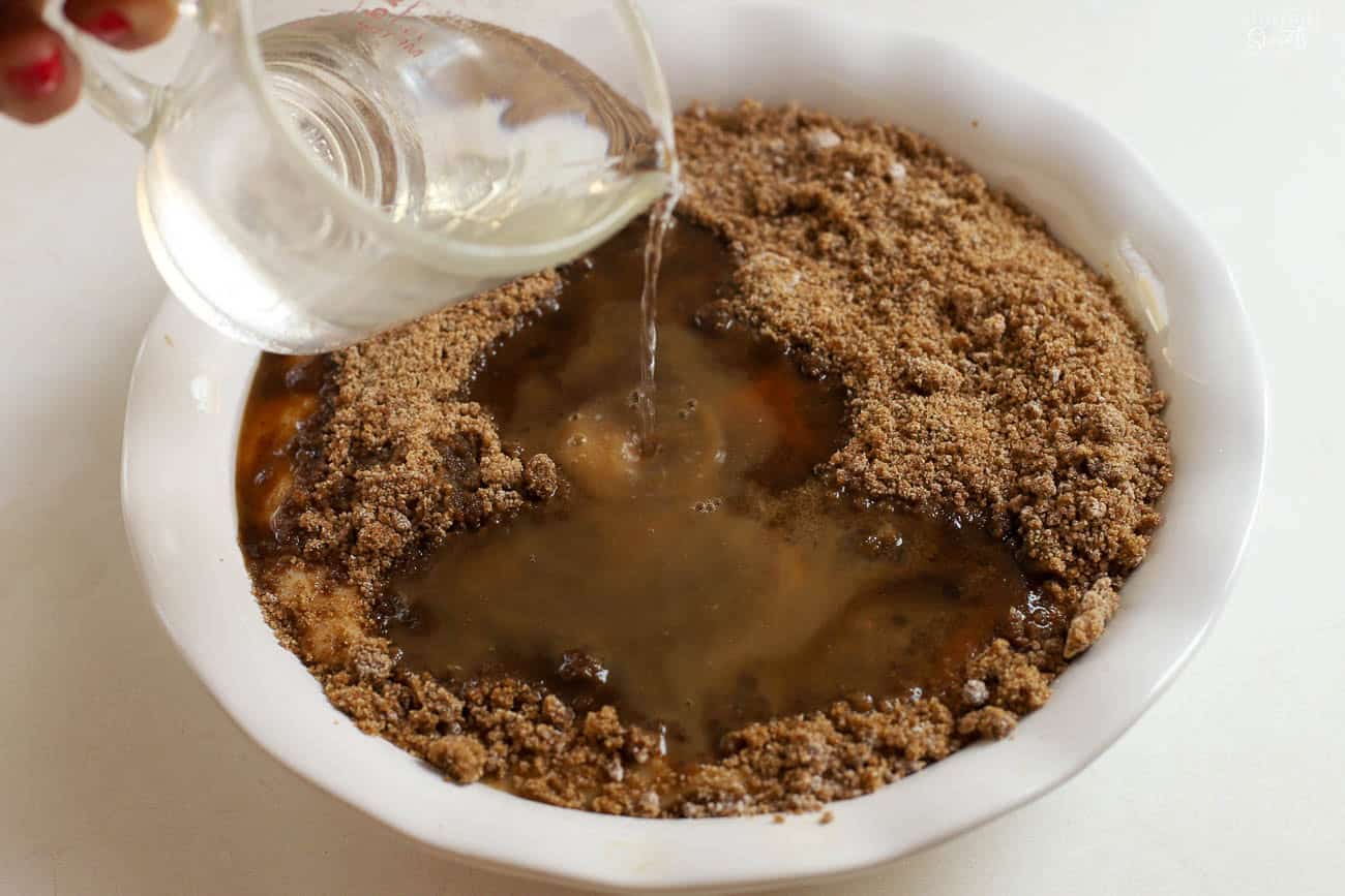 Water being poured over brown sugar and cake batter in a white baking dish.