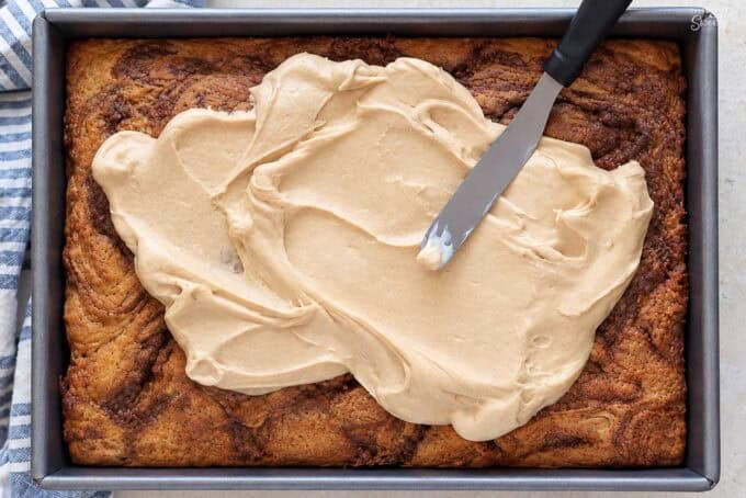 Maple frosting being spread on a maple cinnamon swirl cake in a metal cake pan.