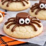 Spider cookies (made from sugar cookies, chocolate frosting, and peanut butter cups) on parchment paper next to an orange napkin.