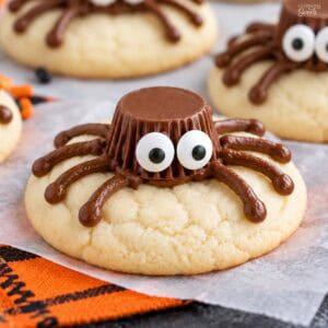 Spider cookies (made from sugar cookies, chocolate frosting, and peanut butter cups) on parchment paper next to an orange napkin.