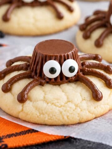 Spider cookies (made from sugar cookies, chocolate frosting, and peanut butter cups) on parchment paper next to an orange napkin.