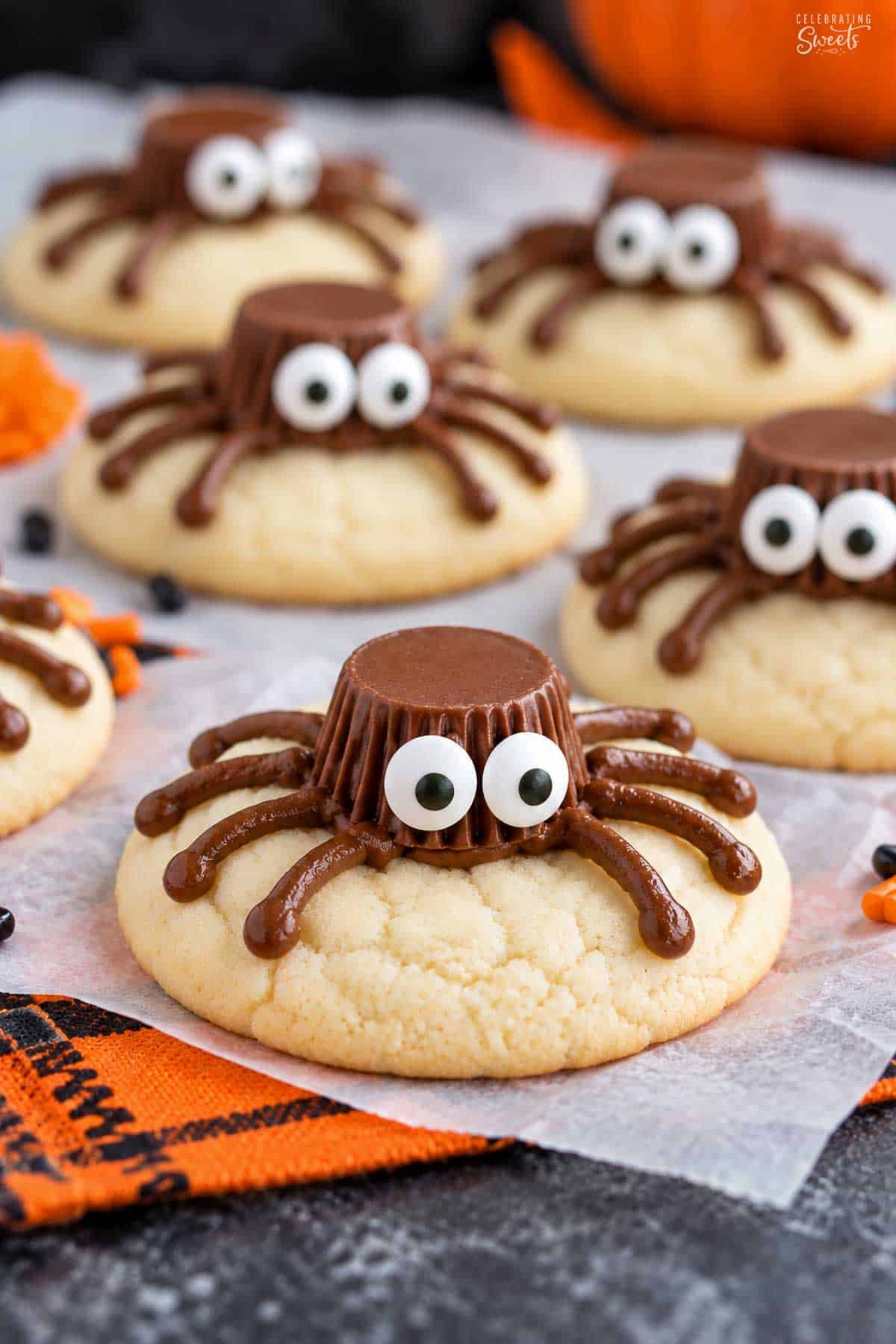 Spider cookies (made from sugar cookies, chocolate frosting, and peanut butter cups) on parchment paper next to an orange napkin.